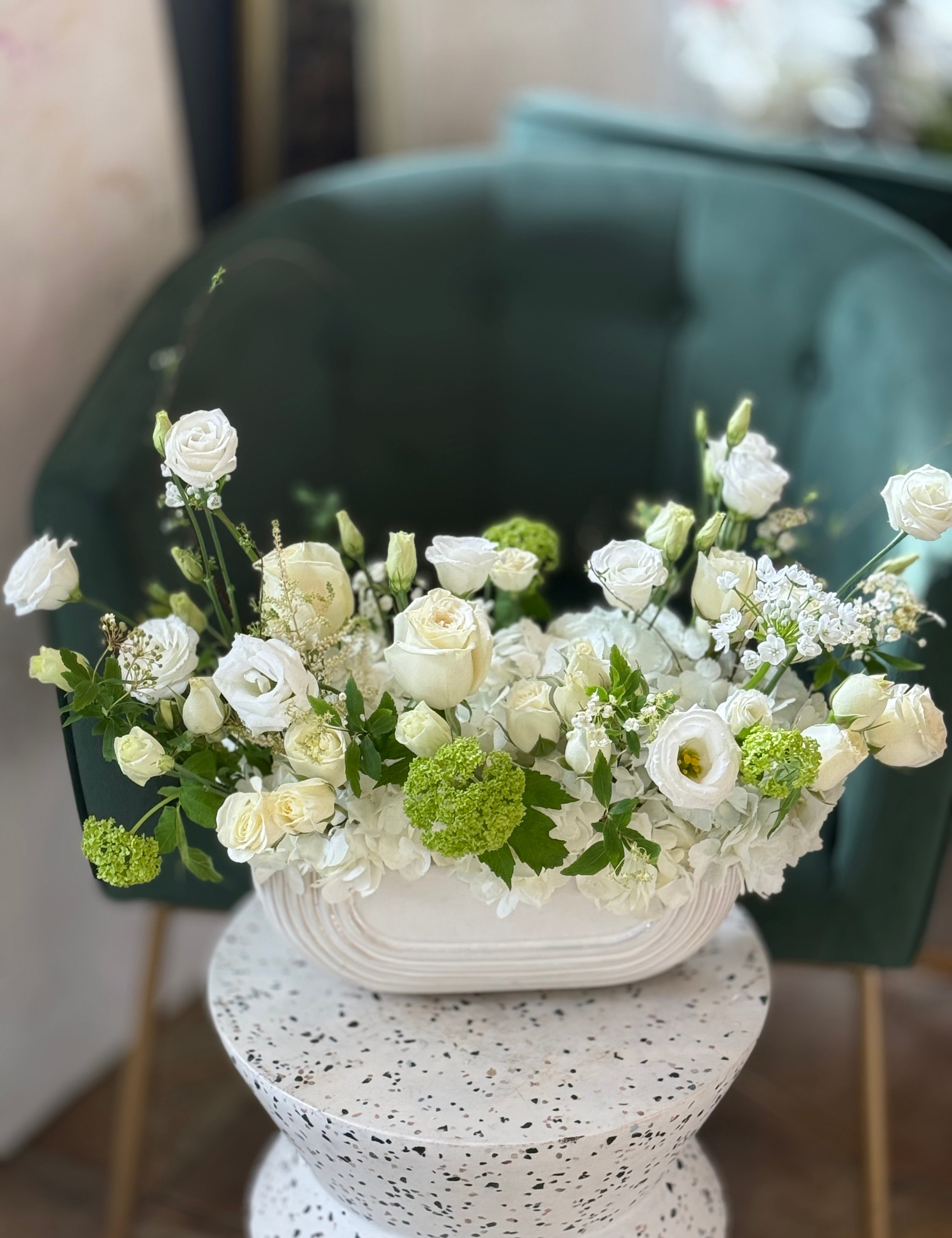 A floral arrangement featuring a variety of flowers including roses and other greenery, arranged in a white container, placed on a white surface with a green chair in the background.