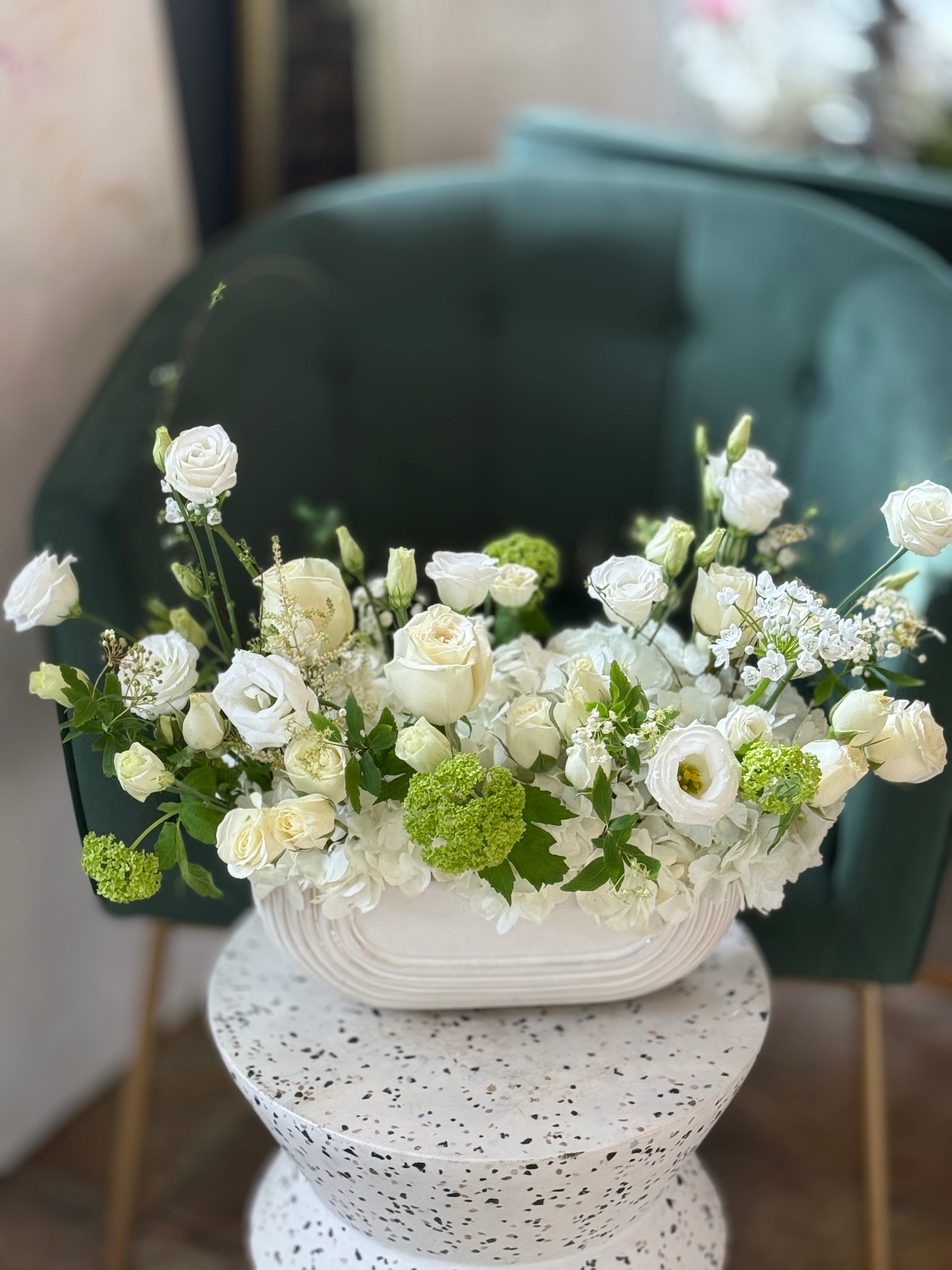 A floral arrangement featuring a variety of flowers including roses and other greenery, arranged in a white container, placed on a white surface with a green chair in the background.