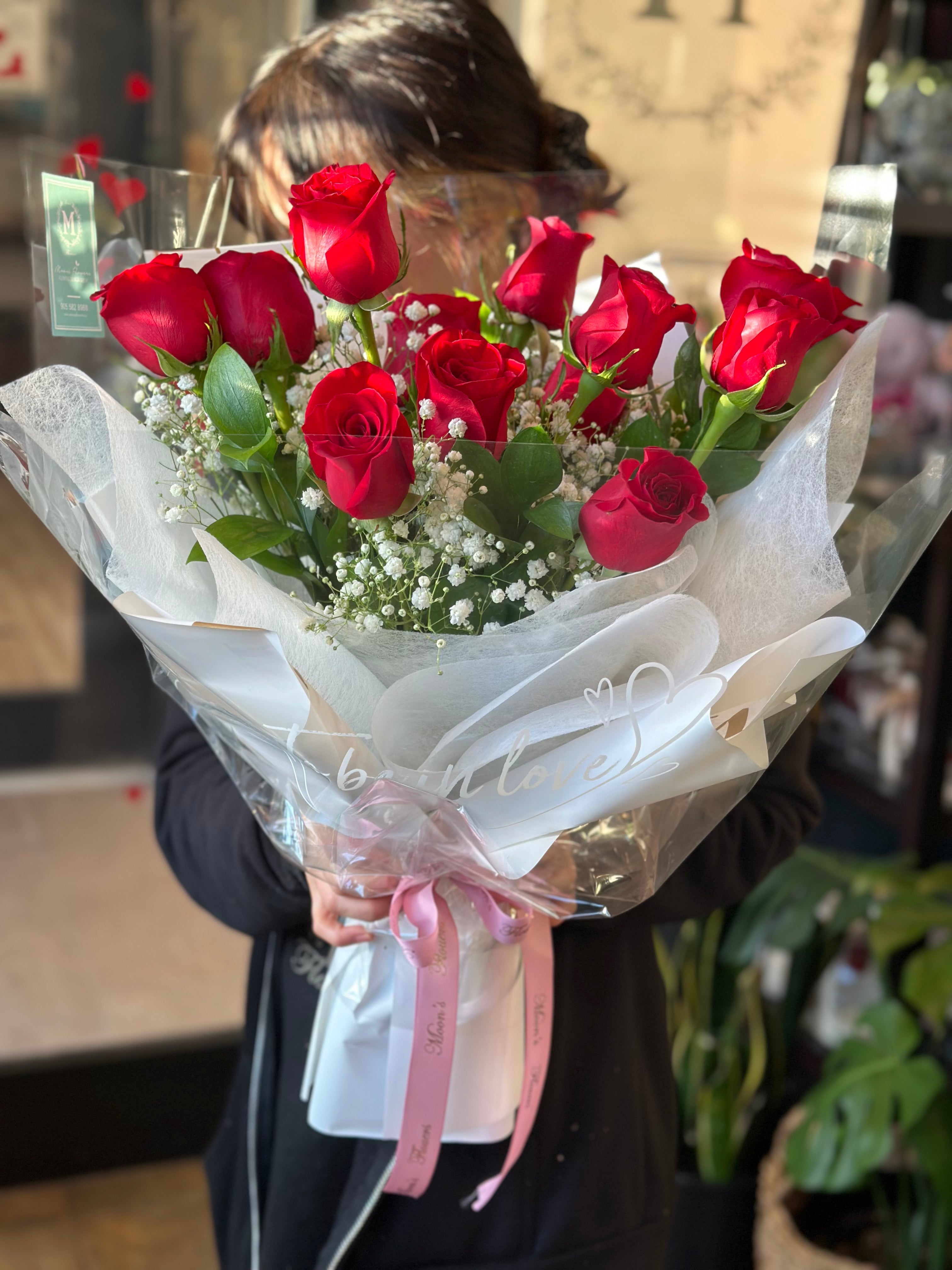 A classic Mother's Day dozen red roses bouquet, beautifully accented with white baby's breath. The arrangement is wrapped in crisp white paper and clear floral film, finished with a delicate pink ribbon. A perfect romantic gift from Moon's Flowers Toronto