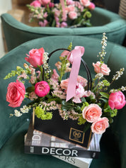 A floral arrangement with peonies, spray roses, lisianthus, and statice in a black box, tied with a pink ribbon, displayed on a stack of magazines.
