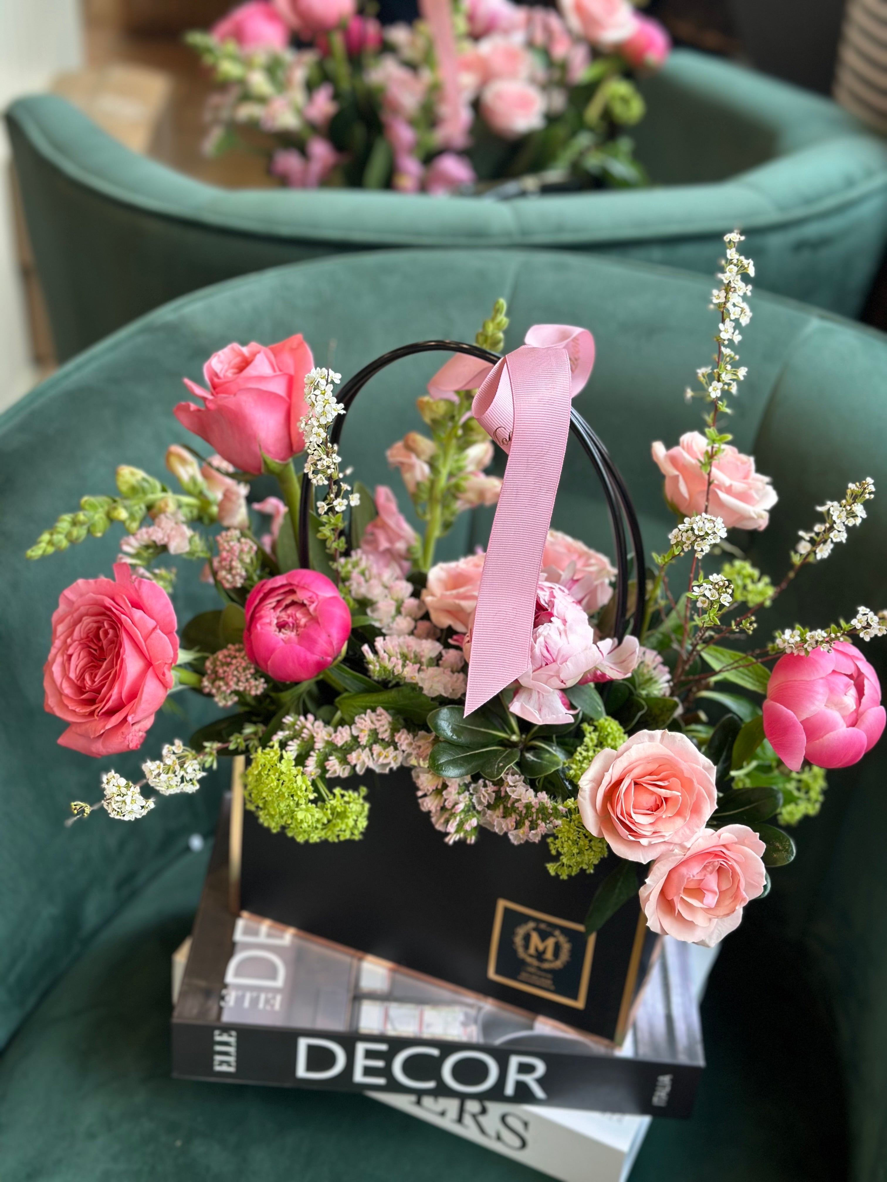 A floral arrangement with peonies, spray roses, lisianthus, and statice in a black box, tied with a pink ribbon, displayed on a stack of magazines.