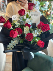 A hand-held Mother's Day bouquet of premium red roses mixed with fresh eucalyptus, wrapped in a modern black textured paper with a signature white brand card. This elegant arrangement by Moon's Flowers Toronto is perfect for a romantic surprise