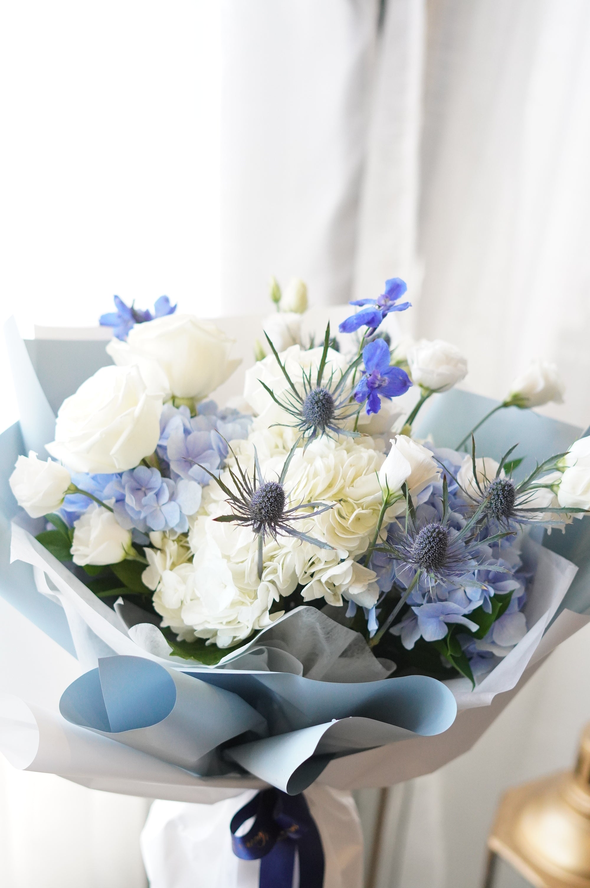 A hand-tied "Frozen Blooms" bouquet wrapped in layered light blue and white decorative paper, featuring white roses, blue hydrangeas, and striking blue thistles.