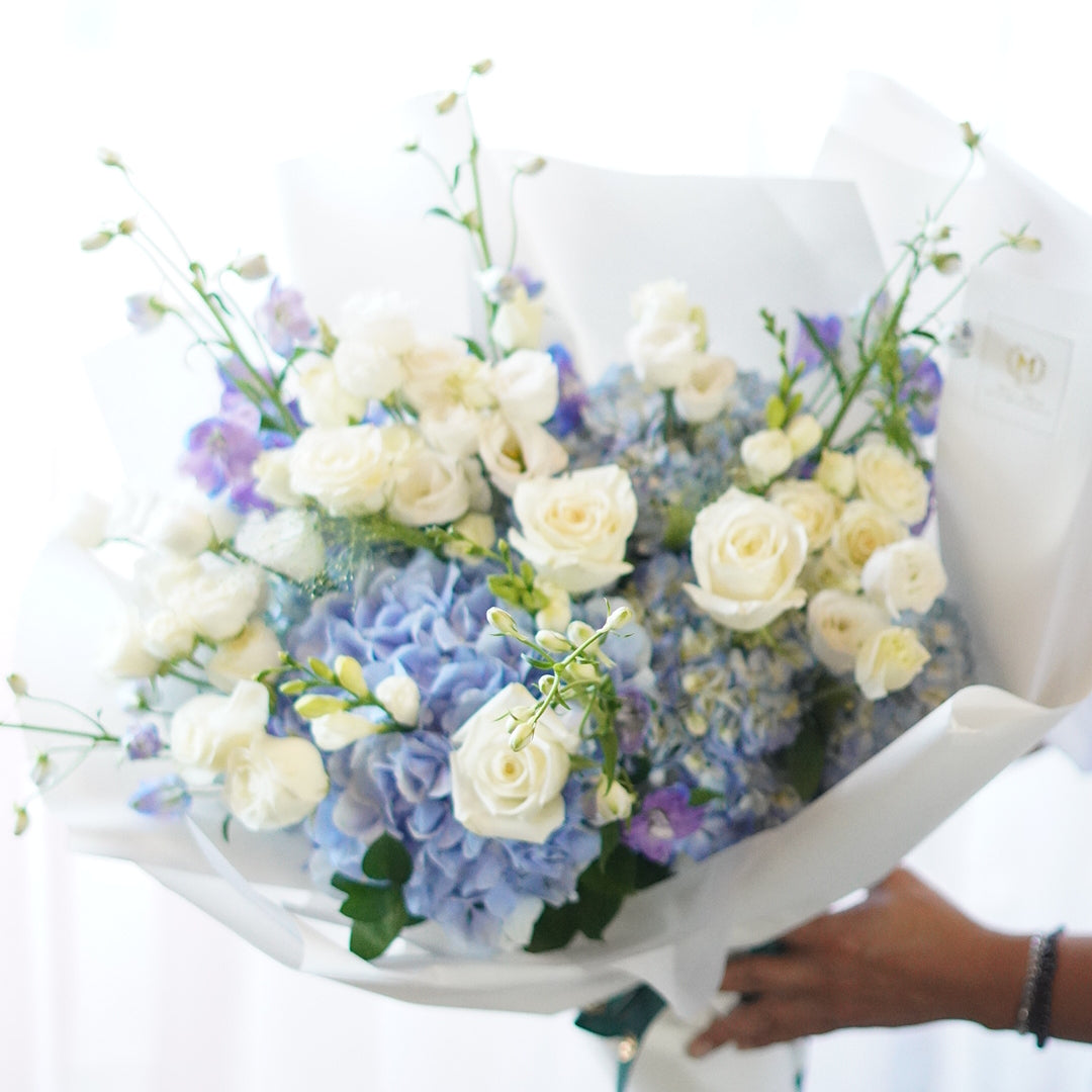 A large floral bouquet featuring light blue hydrangeas, white roses, and delicate blue delphinium accents, wrapped in layered white decorative paper with a dark green ribbon.