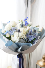 A lush bouquet of white and light blue hydrangeas, white roses, and purple-tipped lisianthus in a clear glass vase
