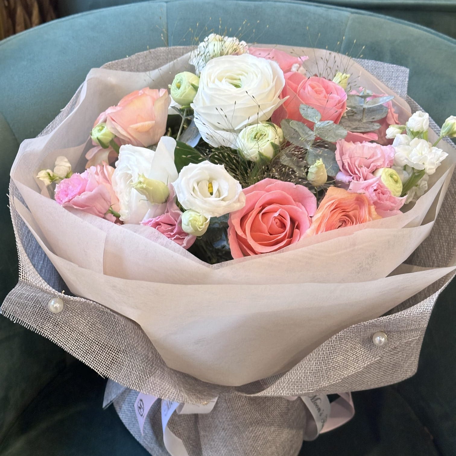 A luxurious Mother's Day flower bouquet featuring soft pink and peach roses, white lisianthus, and fresh eucalyptus leaves, elegantly wrapped in cream and grey textured paper