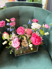 A luxury flower arrangement featuring soft pink peonies, creamy white roses, delicate ranunculus, lisianthus, orchids, and seasonal fillers, presented in a modern clear acrylic box with a natural moss base, displayed on a green upholstered chair.