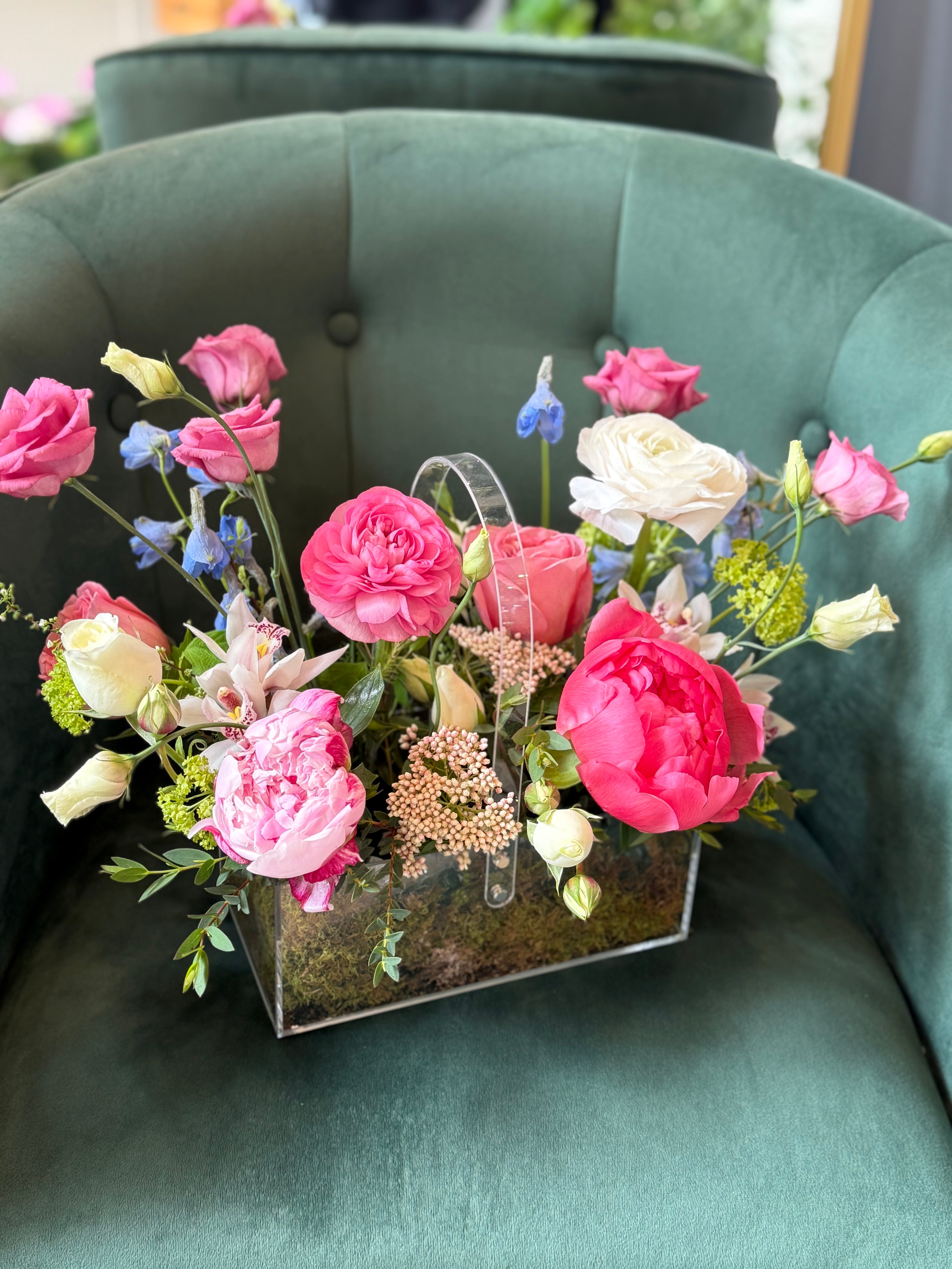 A luxury flower arrangement featuring soft pink peonies, creamy white roses, delicate ranunculus, lisianthus, orchids, and seasonal fillers, presented in a modern clear acrylic box with a natural moss base, displayed on a green upholstered chair.