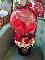 A luxury red rose arrangement featuring a clear crystal-clear balloon with a gold butterfly decal and fairy lights. Set in a black base with white baby's breath  and finished with a satin red bow for mother's day gift in Oakville
