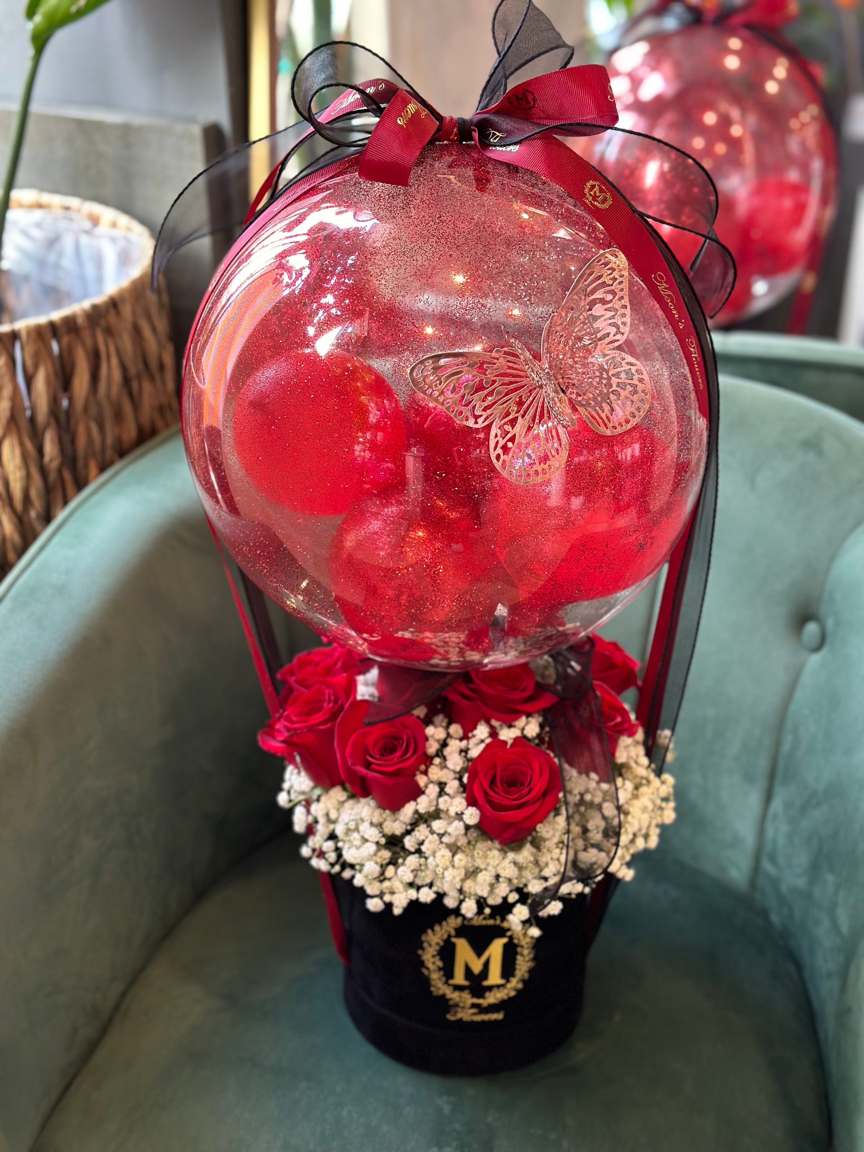 A luxury red rose arrangement featuring a clear crystal-clear balloon with a gold butterfly decal and fairy lights. Set in a black base with white baby's breath  and finished with a satin red bow for mother's day gift in Oakville