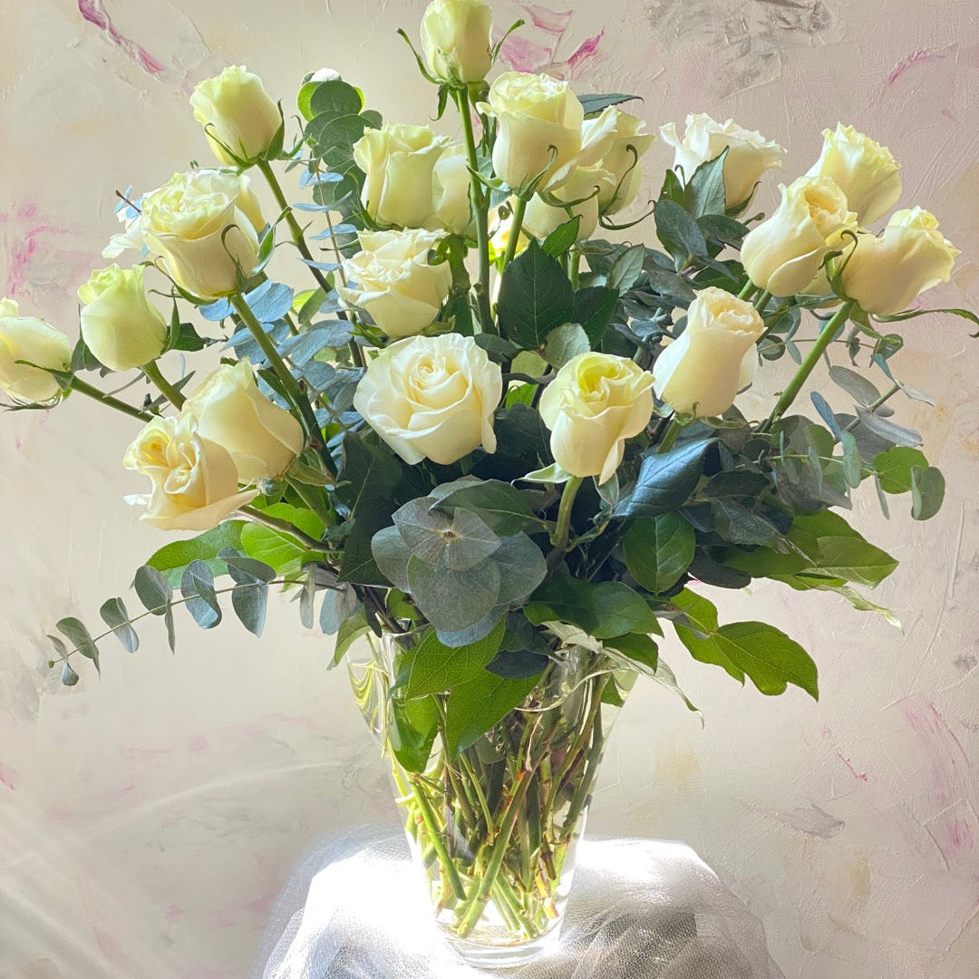 • A sophisticated Mother's Day arrangement of premium white roses with fresh eucalyptus in a clear glass vase.
• The bouquet is presented on a white draped stand, showcasing an elegant and pure floral design by Moon's Flowers Toronto.