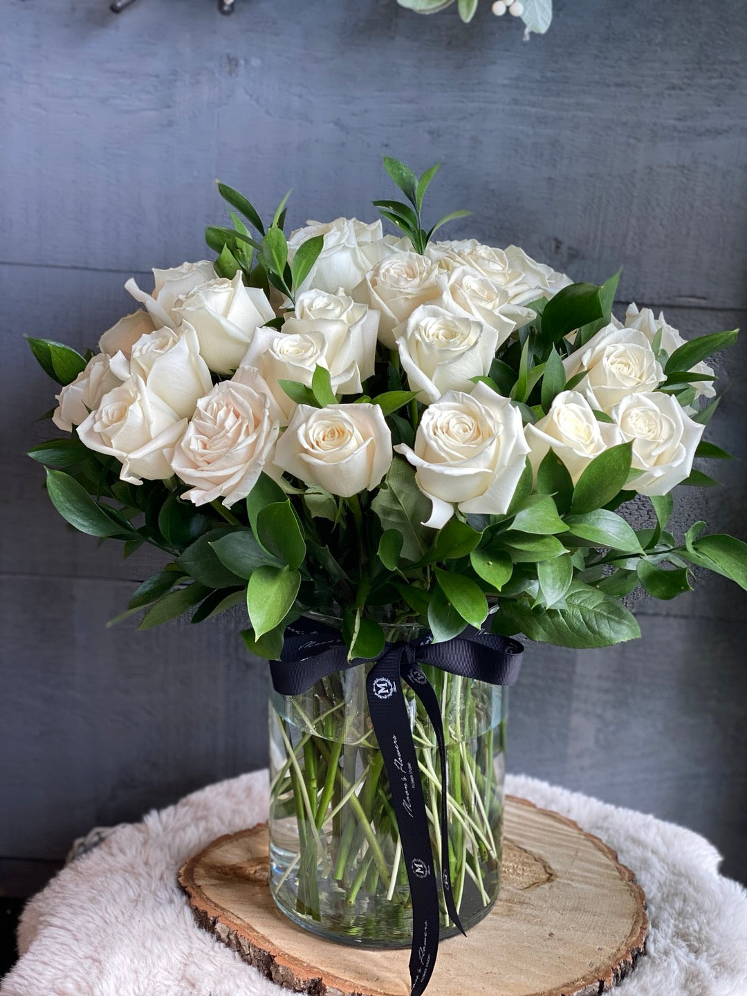 • A stunning Mother's Day bouquet of white roses and lush greenery in a clear glass vase tied with a signature Moon's Flowers black ribbon.
• The arrangement is placed on a rustic wood slice and a white faux fur rug, creating a cozy and elegant atmosphere for home delivery in Toronto.