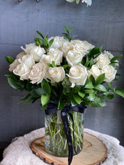 • A stunning Mother's Day bouquet of white roses and lush greenery in a clear glass vase tied with a signature Moon's Flowers black ribbon.
• The arrangement is placed on a rustic wood slice and a white faux fur rug, creating a cozy and elegant atmosphere for home delivery in Toronto.