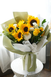 A vibrant bouquet featuring large yellow sunflowers, cream-colored roses, and white carnations, elegantly wrapped in layers of sage green and translucent white paper, tied with a matching green ribbon.