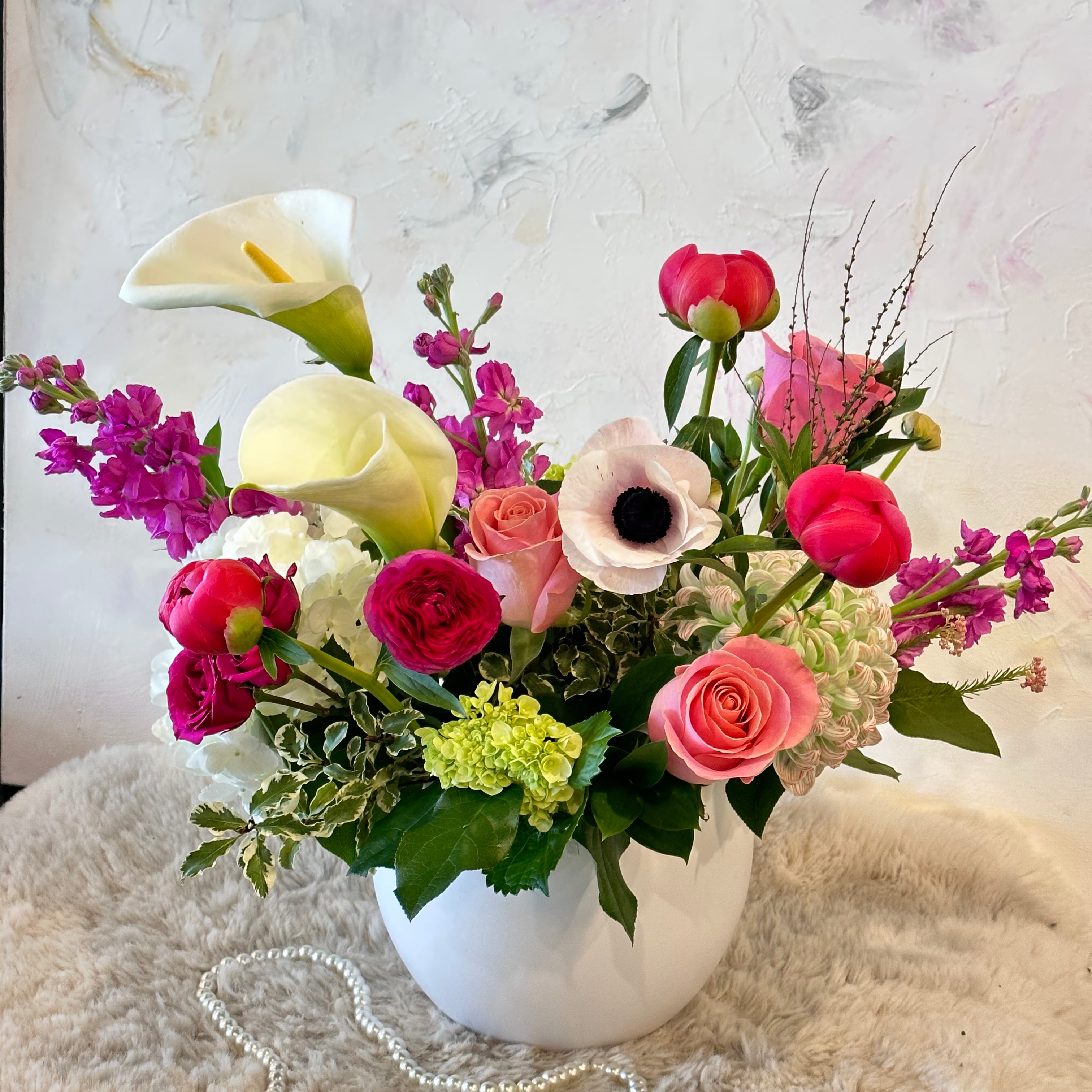 A vibrant floral arrangement in a white ceramic vase featuring white Calla Lilies, hot pink roses, purple stock flowers, and green hydrangeas. The arrangement is set against a soft white faux fur rug and a pearl necklace on a textured background.