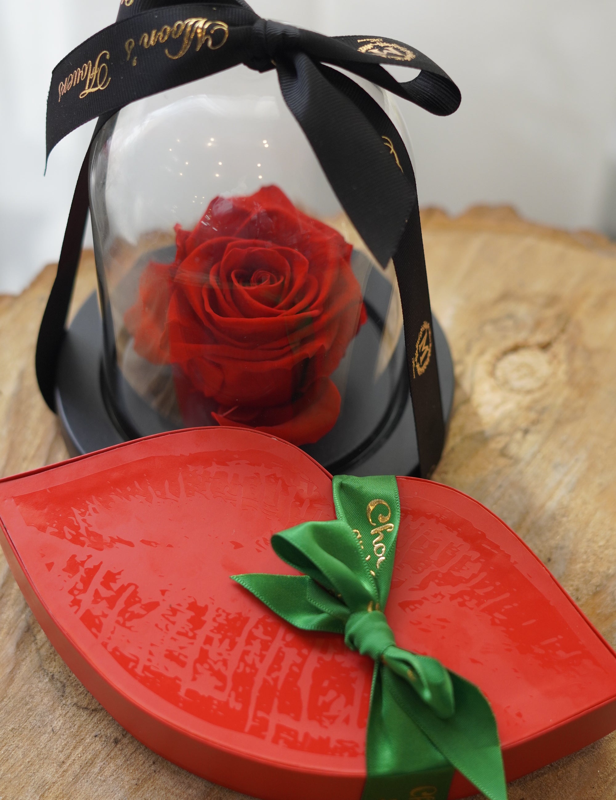 Lip-shaped red gift box with a green ribbon and a preserved rose inside, on a wooden surface.