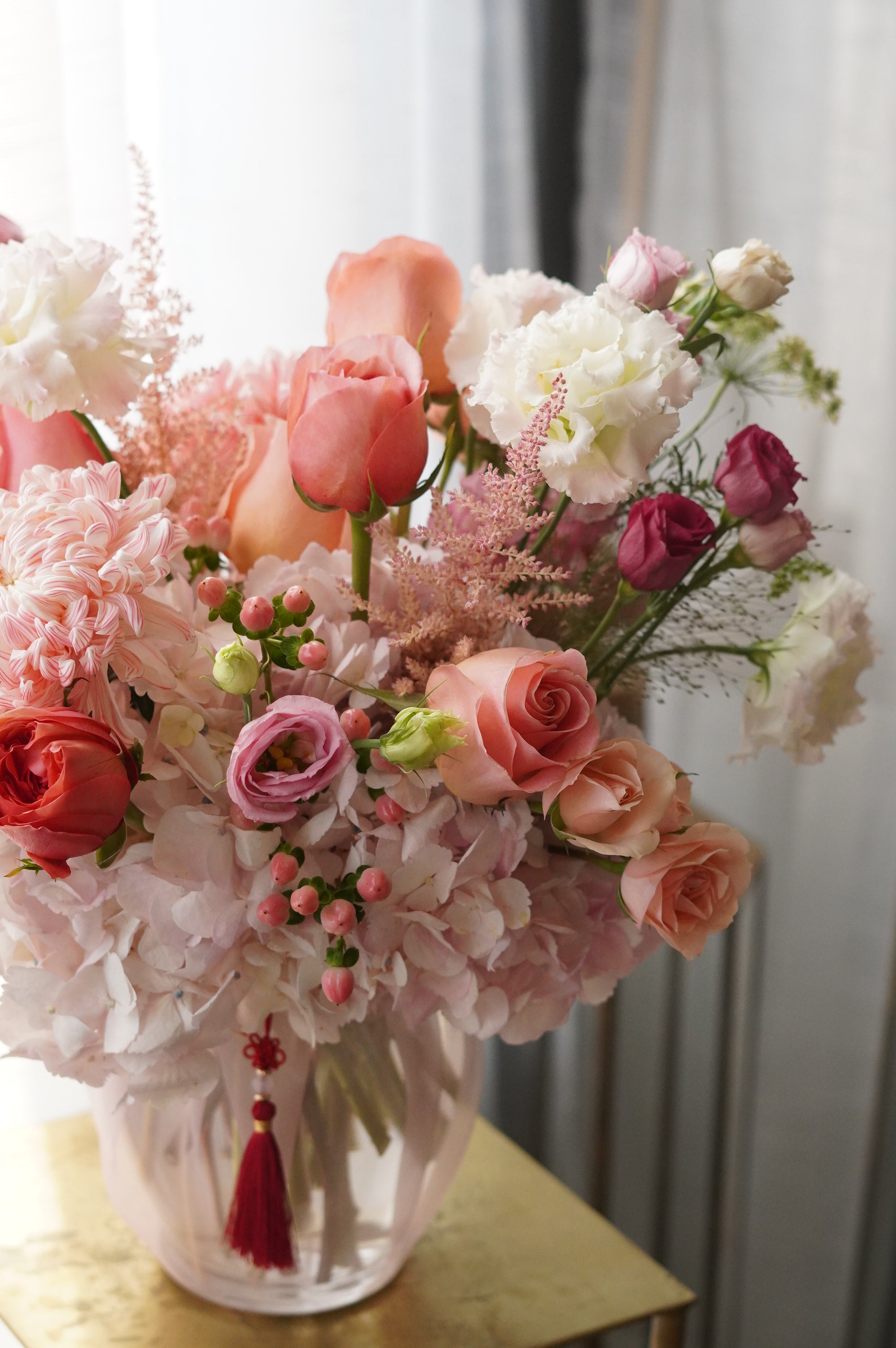 Close-up of a Mother's Day floral arrangement in a glass vase featuring peach roses, pink spray roses, and dried textures with a traditional red Korean tassel. Luxury flower delivery in Toronto and Oakville