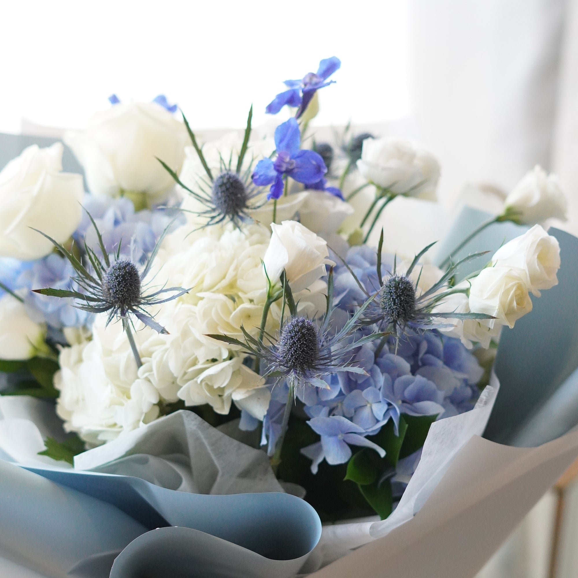 Close-up of a floral arrangement featuring large clusters of serene blue and pure white hydrangeas, accented with elegant white roses and lisianthus, presented in a cylindrical glass vase with clear water.