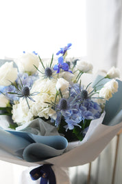 Close-up of a floral arrangement featuring large clusters of serene blue and pure white hydrangeas, accented with elegant white roses and lisianthus, presented in a cylindrical glass vase with clear water.