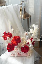 Red preserved roses and dried white flowers in a clear gift box.