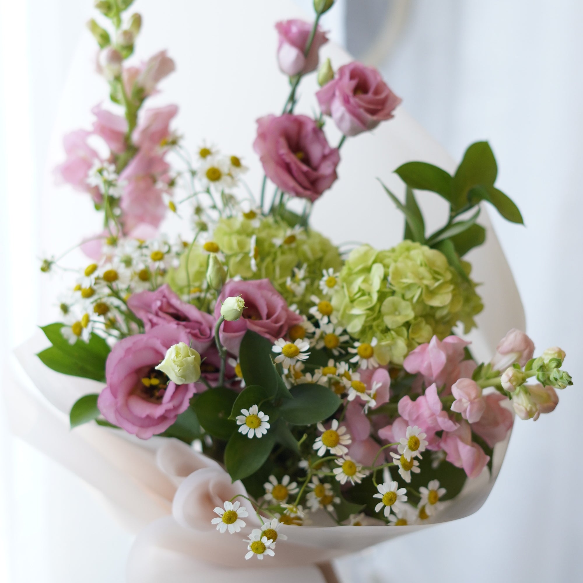 Whimsical summer flower bouquet with pink lisianthus, matricaria daisies, and green hydrangeas wrapped in soft blush pink paper.