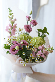 Whimsical summer flower bouquet with pink lisianthus, matricaria daisies, and green hydrangeas wrapped in soft blush pink paper.