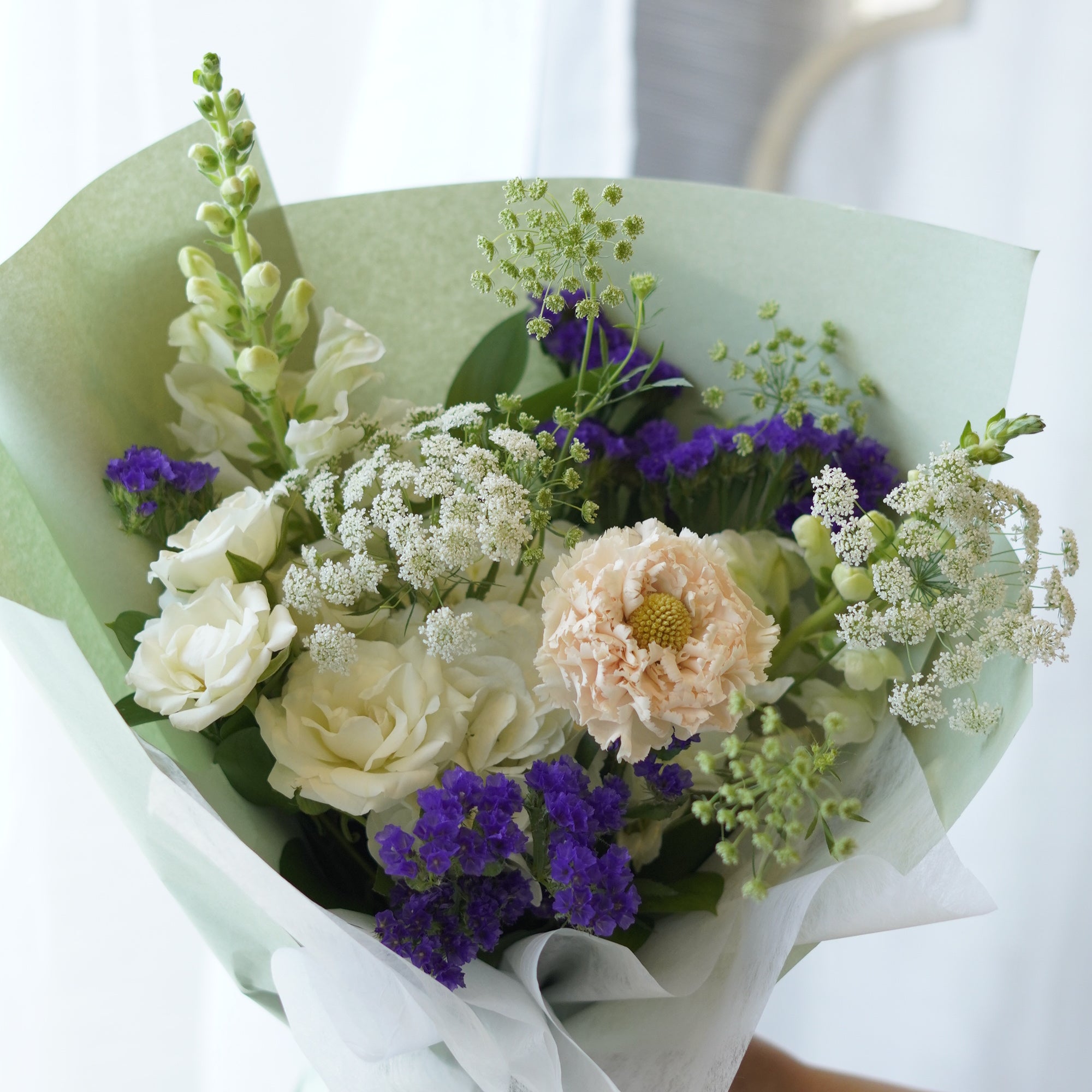 Fresh summer flower bouquet featuring purple statice, white roses, and snapdragons wrapped in sage green paper by a local florist in Oakville
