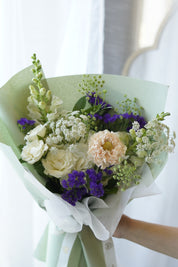 Fresh summer flower bouquet featuring purple statice, white roses, and snapdragons wrapped in sage green paper by a local florist in Oakville