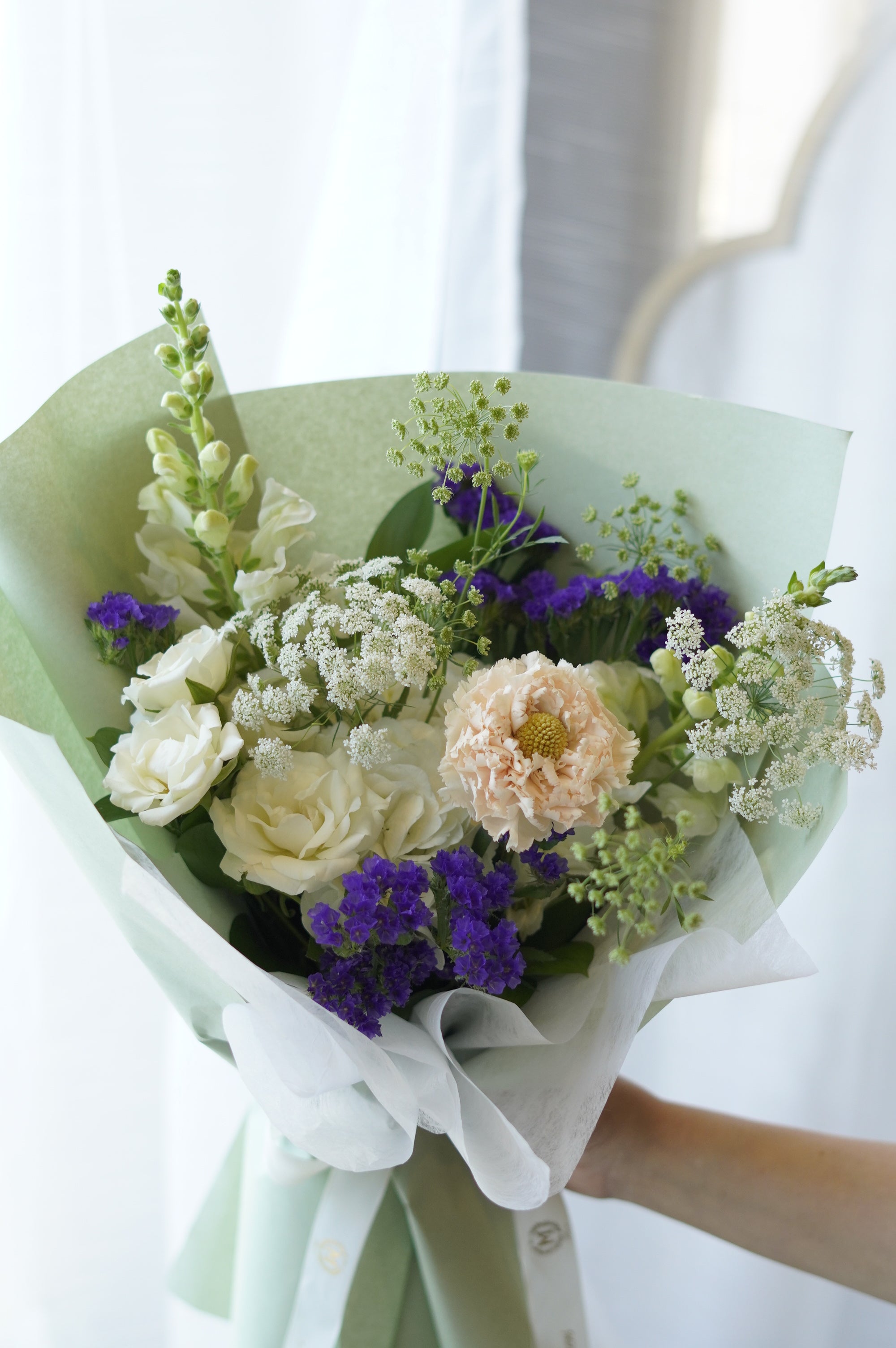 Fresh summer flower bouquet featuring purple statice, white roses, and snapdragons wrapped in sage green paper by a local florist in Oakville
