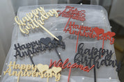 Assorted acrylic cake toppers including Happy Birthday, Happy Anniversary, Feliz Aniversario, and Welcome styles in multiple colors on white tulle