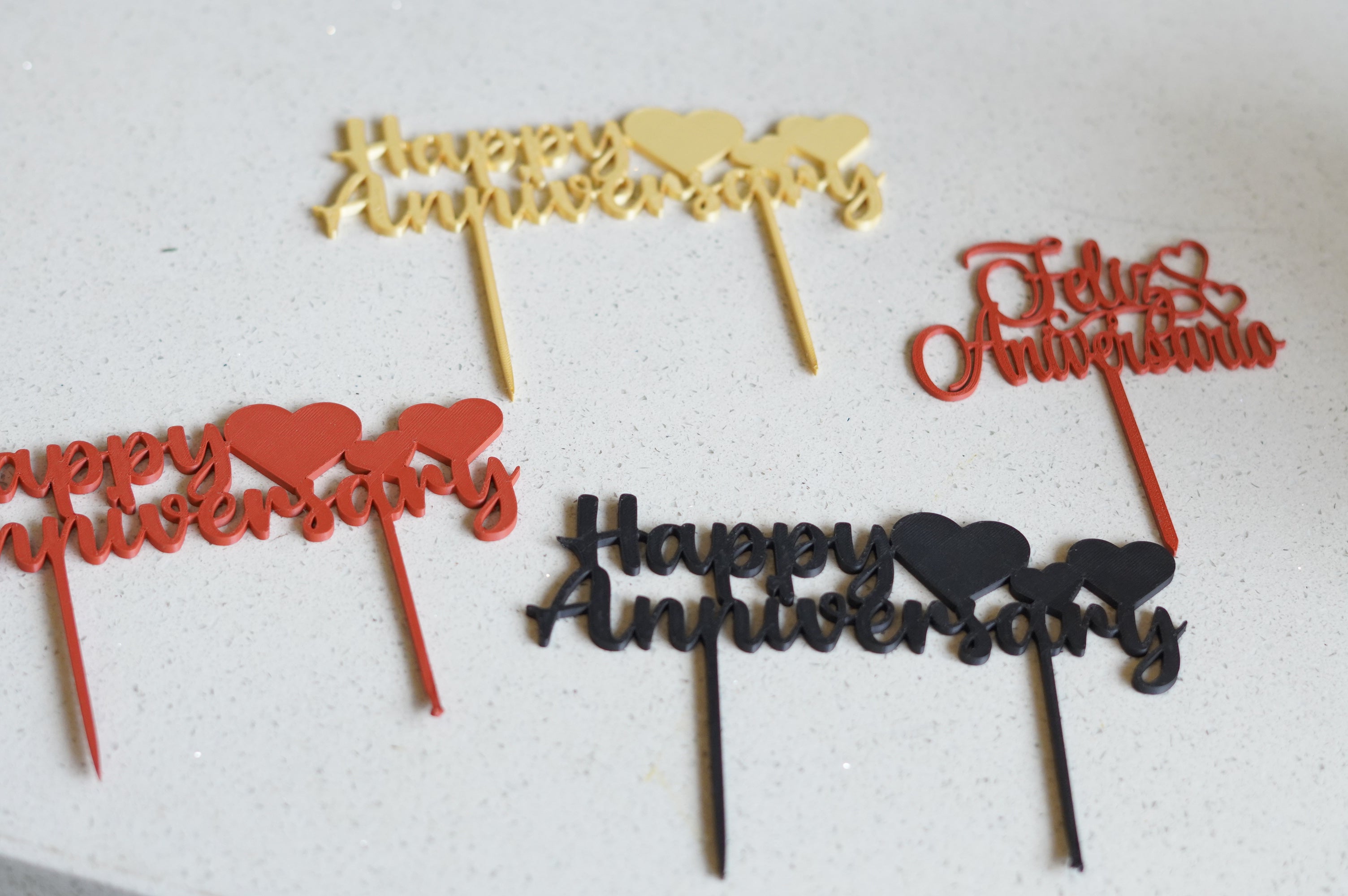 Gold, red, and black "Happy Anniversary" and "Feliz Aniversario" acrylic cake toppers on white surface