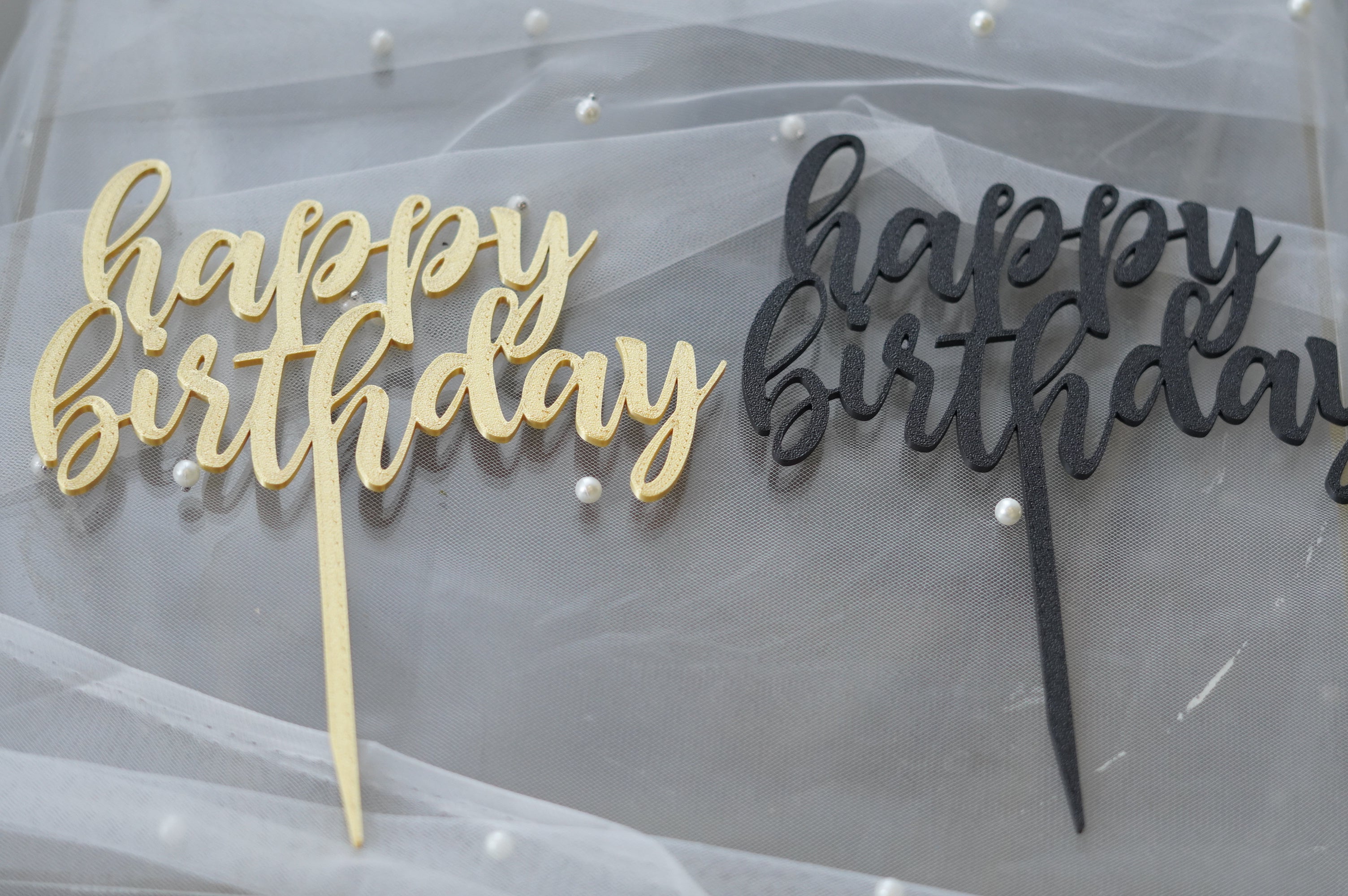  Gold and black "happy birthday" acrylic cake toppers laid on white tulle with pearl accents
