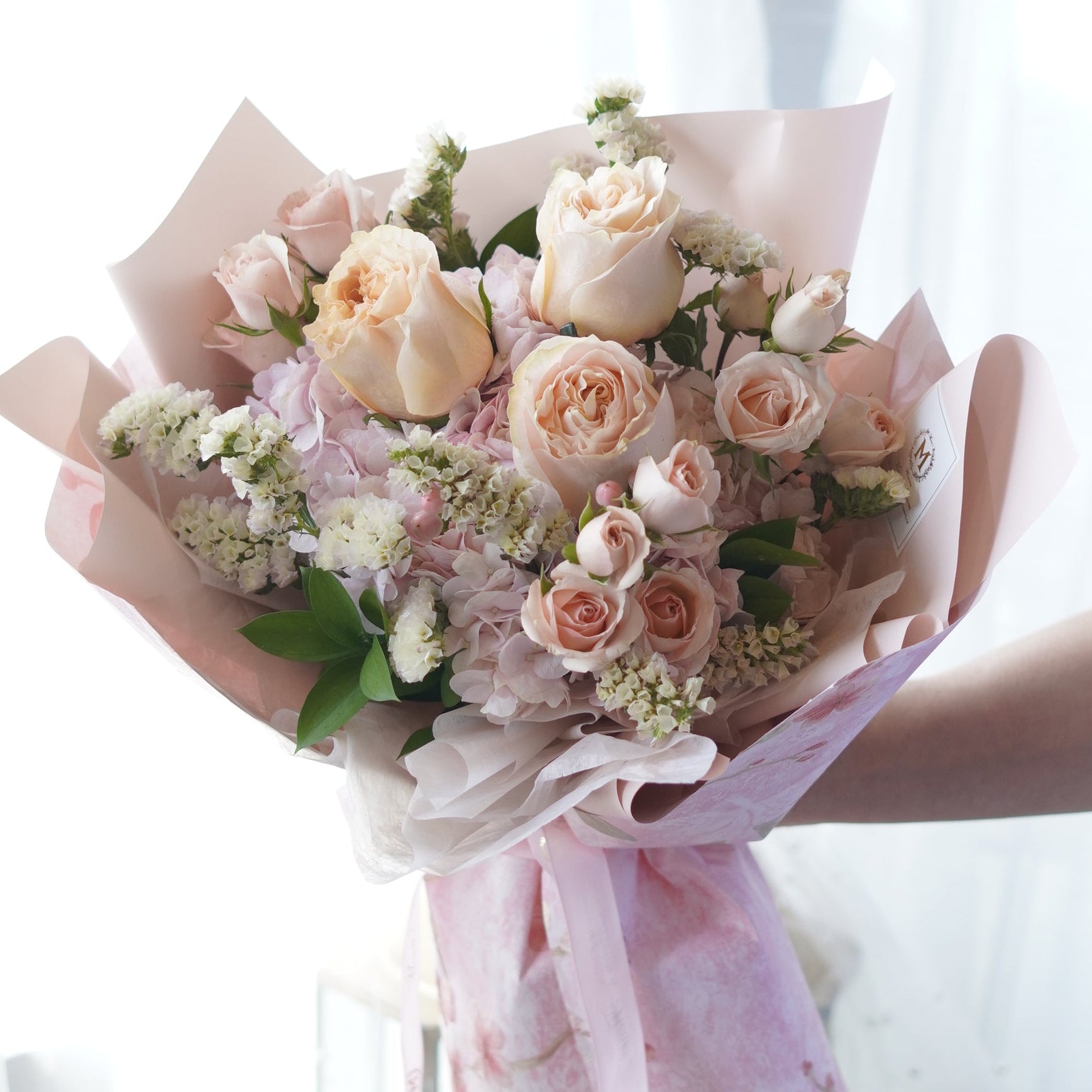 **Handheld Pink Blossom bouquet featuring peach roses, pink spray roses, light purple hydrangeas, and white fillers wrapped in layered pink floral wrapping paper with a ribbon.**
