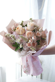 **Handheld Pink Blossom bouquet featuring peach roses, pink spray roses, light purple hydrangeas, and white fillers wrapped in layered pink floral wrapping paper with a ribbon.**
