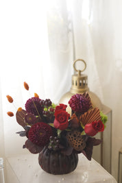 Burgundy dahlias, red roses, lotus seed pod, and orange dried grasses in a ribbed pumpkin-shaped vase