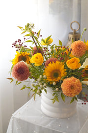 Yellow ranunculus and sunflowers with peach dahlias and greenery in a white vase