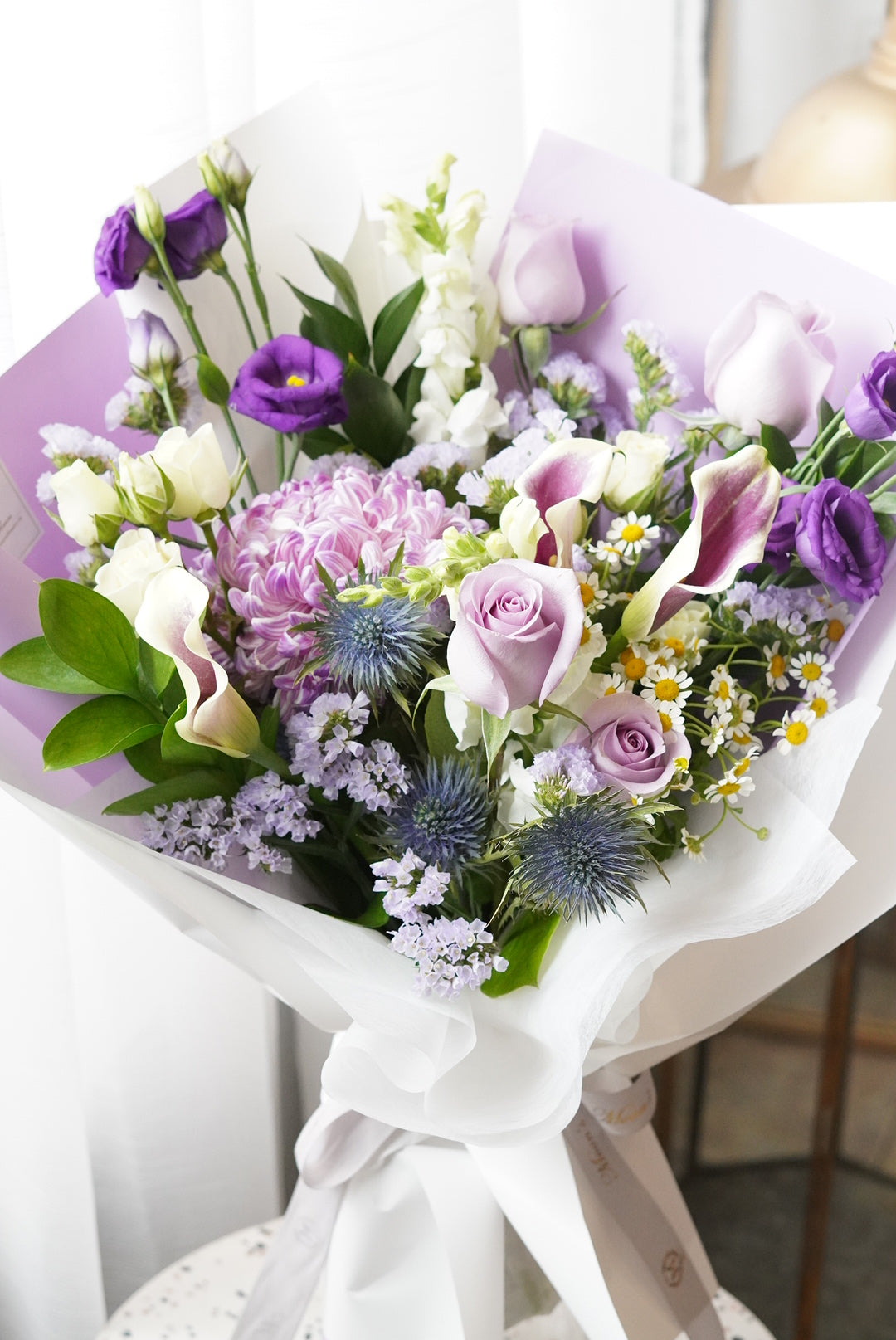 Elegant Purple Rush Mother's Day bouquet featuring lavender roses, purple calla lilies, and blue thistles in a white designer wrap. Luxury hand-tied floral gift by Moon's Flowers, available for same-day delivery in Toronto