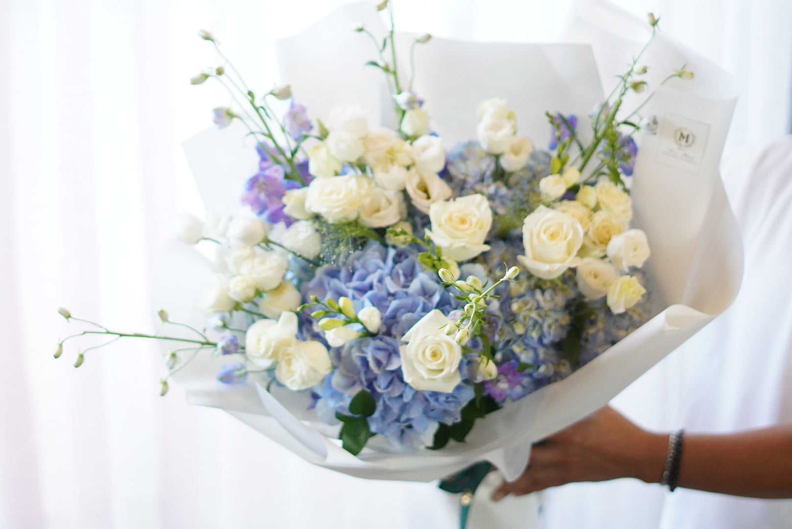 Elegant blue hydrangea and white rose bouquet wrapped in white paper.