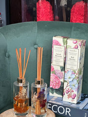 A fragrance diffuser with reeds in a clear glass bottle, placed on a wooden surface, with a floral box in the background.