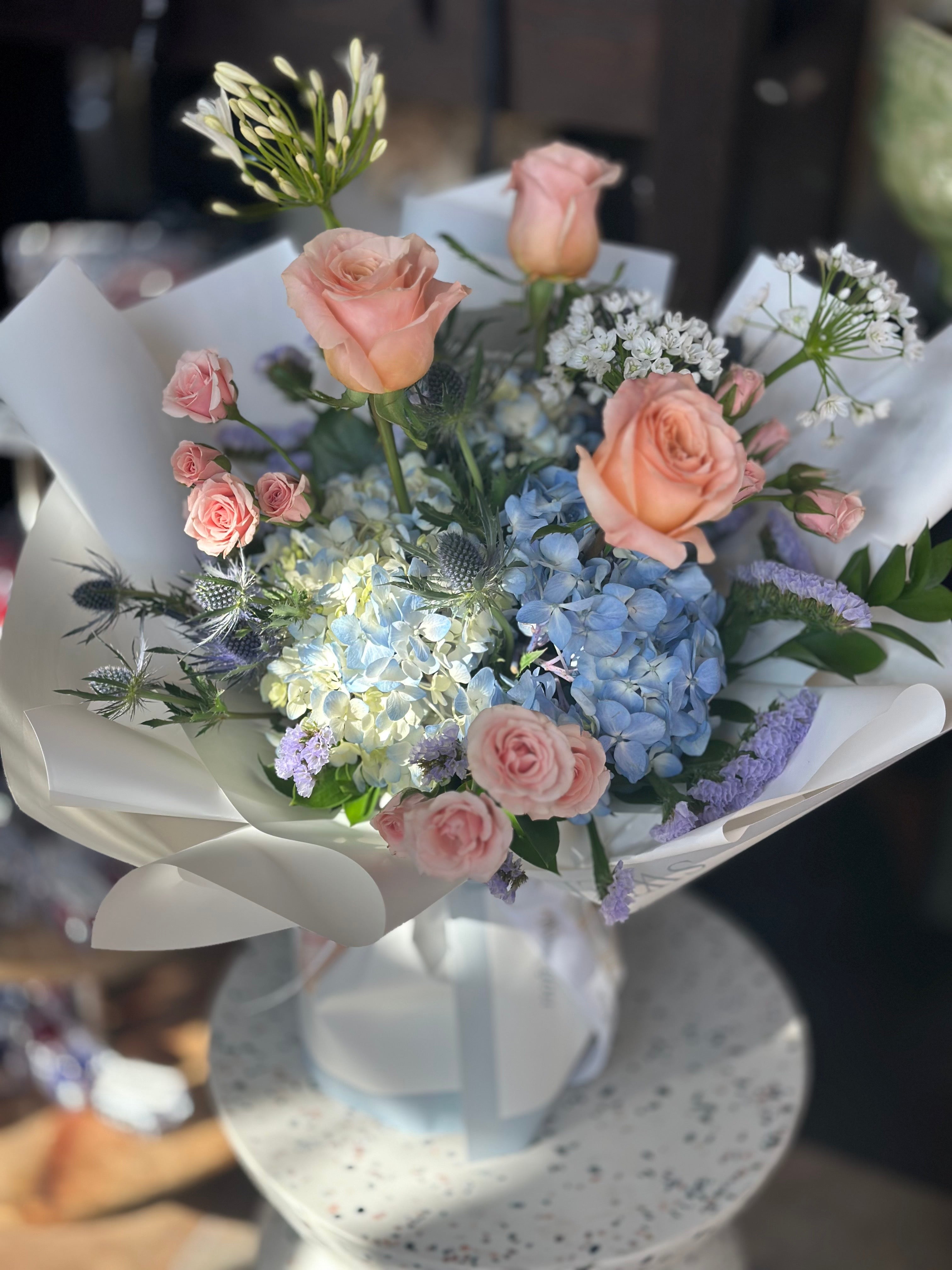 A bouquet of flowers including Hydrangeas, Roses, Statice, Agapanthus, and Thistles with a dreamy color palette of blues, pinks, and champagnes.