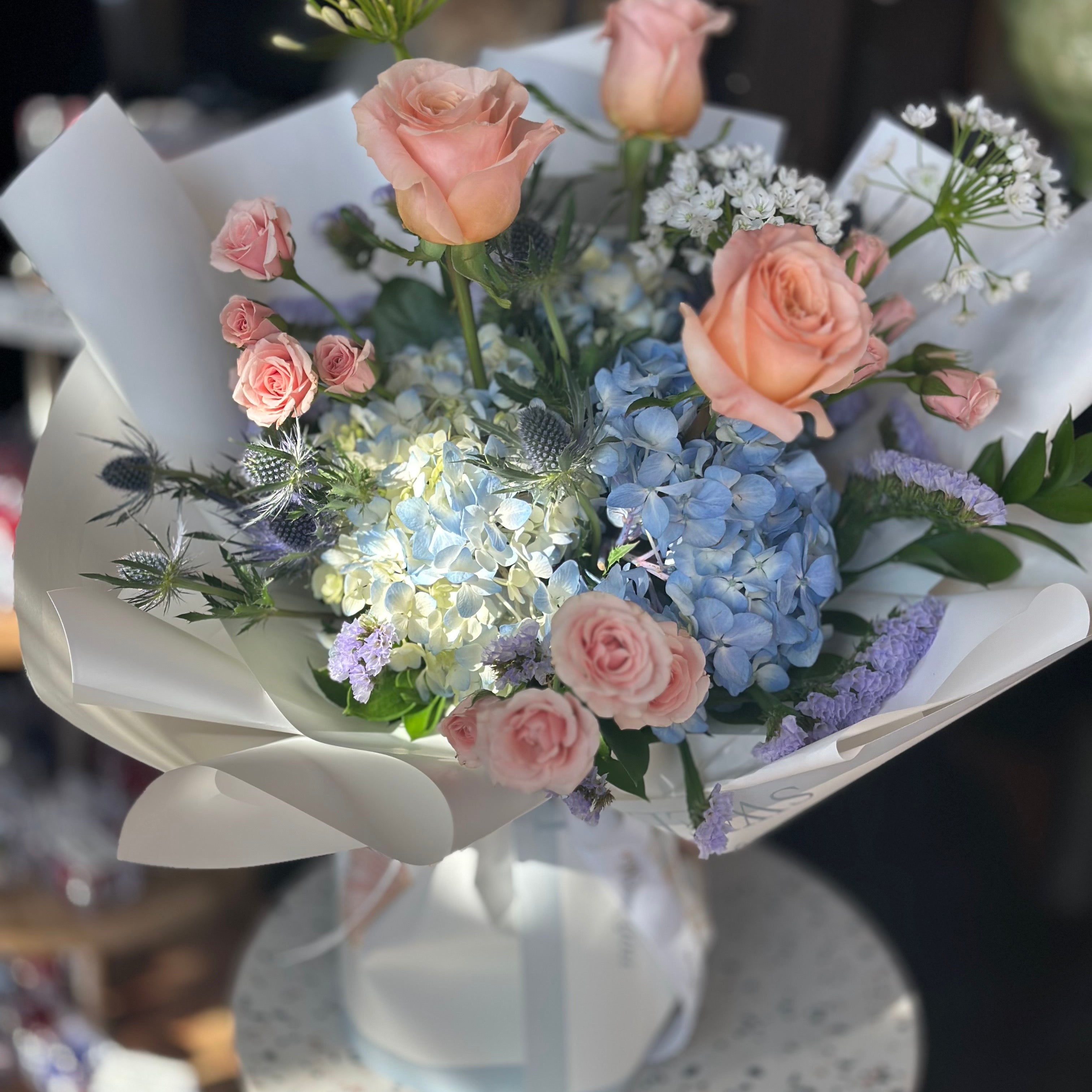 Golden Hour Mother's Day bouquet featuring peach roses, blue hydrangeas, and white ranunculus in a designer wrap. Premium flower delivery in Toronto and Oakville by Moon's Flowers.