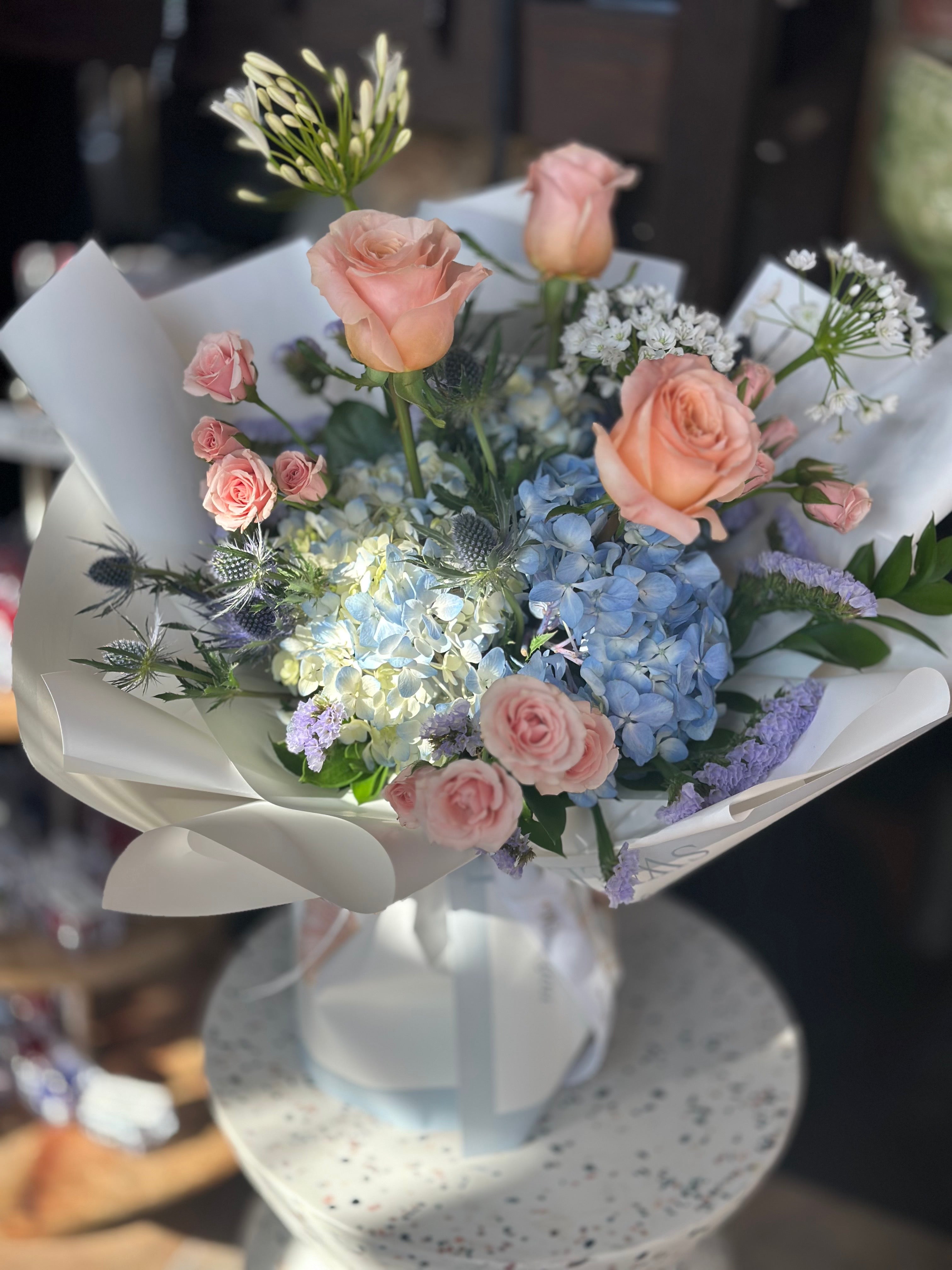 Golden Hour Mother's Day bouquet featuring peach roses, blue hydrangeas, and white ranunculus in a designer wrap. Premium flower delivery in Toronto and Oakville by Moon's Flowers.