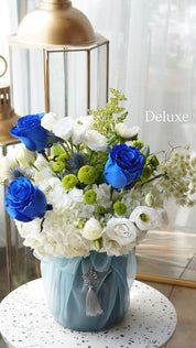 Decorative flower arrangement with blue, white, and green flowers in a light blue vase on a white surface.