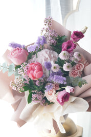 Lush Mother's Day hand-tied bouquet featuring coral and pink peonies, purple lisianthus, and lavender stock flowers wrapped in elegant blush paper.