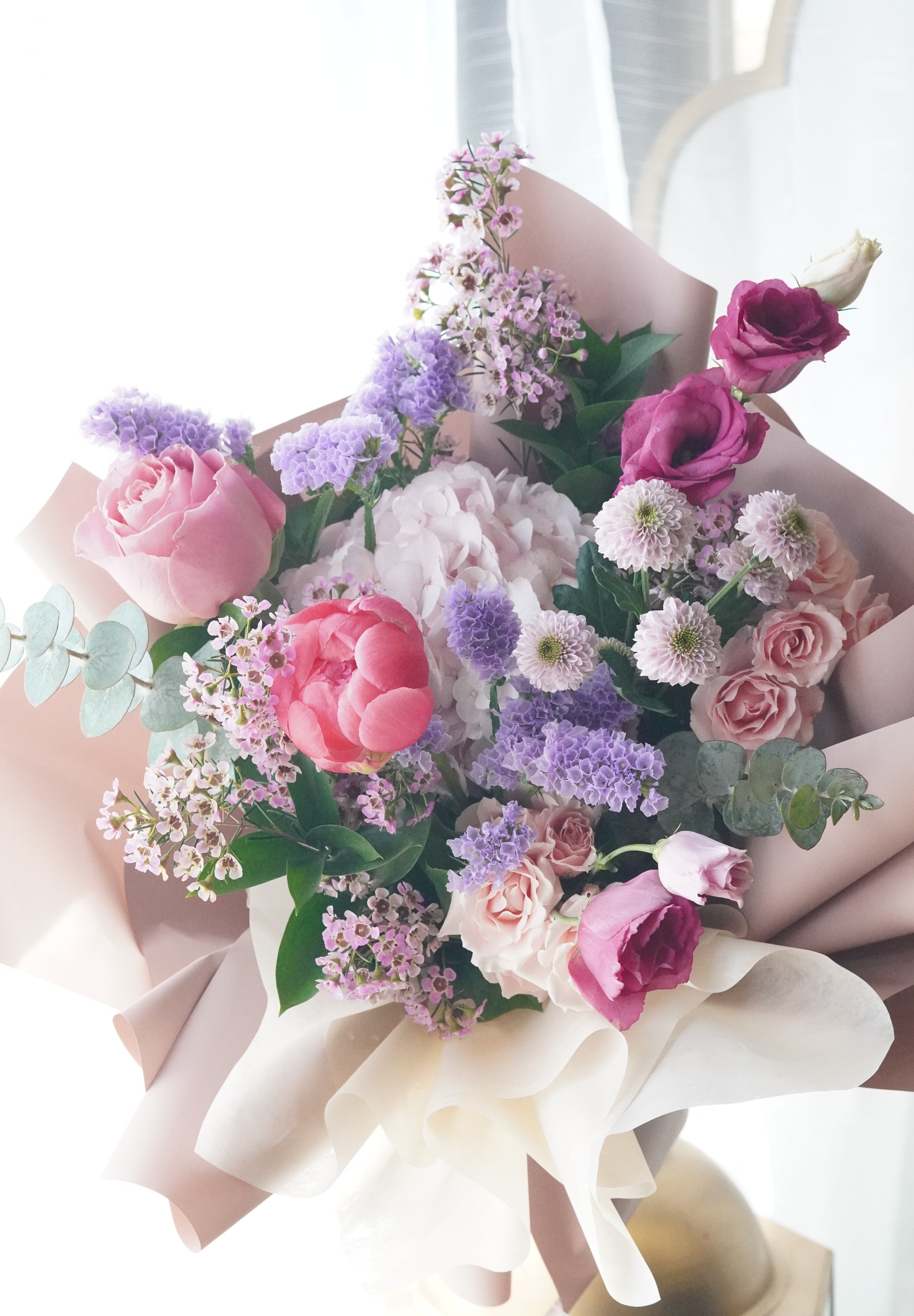 Lush Mother's Day hand-tied bouquet featuring coral and pink peonies, purple lisianthus, and lavender stock flowers wrapped in elegant blush paper.