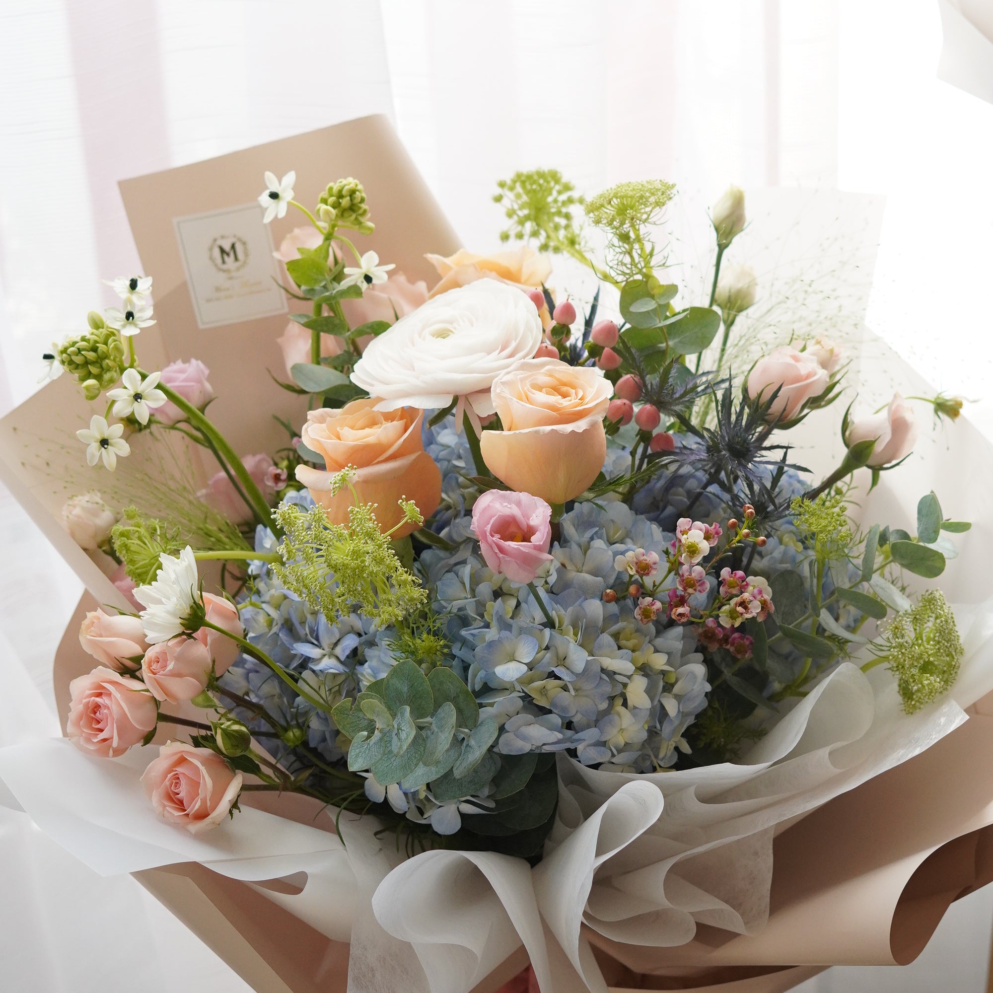 Main view of the Golden Hour bouquet featuring vibrant peach roses, soft blue hydrangeas, and white ranunculus in a luxury designer wrap. Premium Mother's Day flower arrangement by Moon's Flowers Toronto
