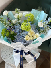 A hand-tied bouquet with a variety of flowers including roses and hydrangeas, wrapped in a signature style with blue and white ribbons and a water bag.