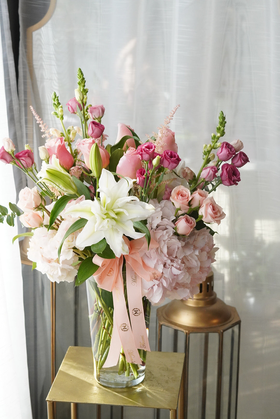 This image showcase the Mix Sympathy Flowers Vase in its Deluxe version. White flowers in this arrangement include lilies, hydrangeas and spray roses. Pink flowers shown are hydrangeas, spray roses, and other seasonal flowers.
