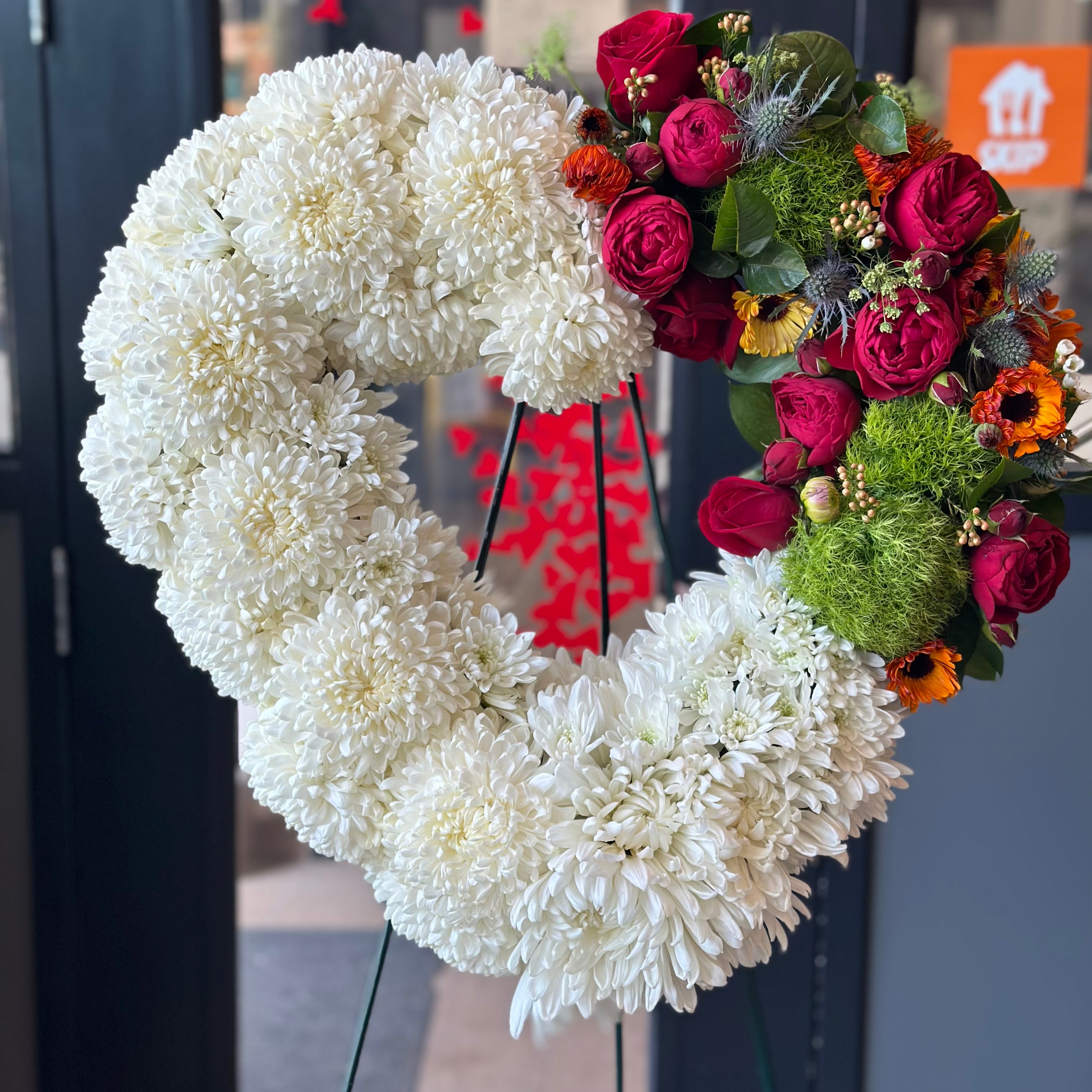 Modern heart-shaped funeral wreath with white carnations and a red rose accent on a standing easel.