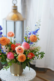 Modern spring floral centerpiece featuring vibrant orange and blue flowers in a sleek gold vase. Elegant 'Designer's Choice' bouquet styled on a terrazzo table, perfect for luxury home decor and Mother's Day gifting